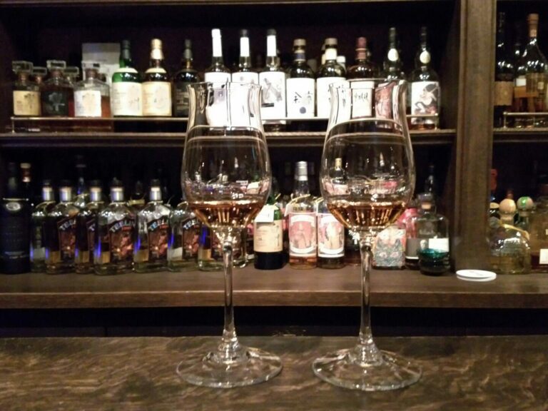 A glass of amber whisky resting on a rich wooden bar counter, with dimly lit shelves of liquor bottles visible in the soft background.
