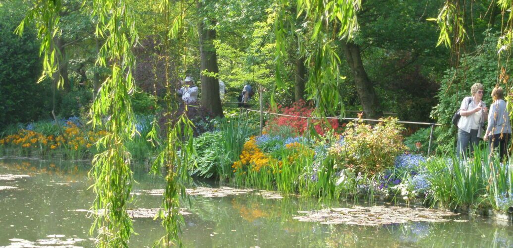 A serene pond with water lilies framed by cascading weeping willow branches, with passersby walking along a garden path in the background.